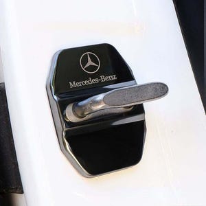 May include: Black car door latch cover with the Mercedes-Benz logo and text. The cover is rectangular with rounded edges and a silver metal latch. The background is white.