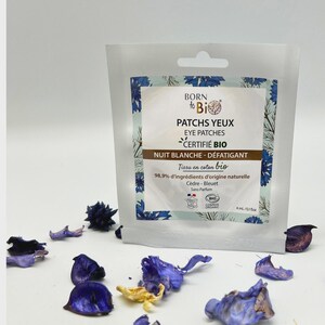 Peut inclure: Un sachet blanc de patchs pour les yeux "BORN to Bio" avec le texte "PATCHS YEUX EYE PATCHES CERTIFIÉ BIO NUIT BLANCHE - DÉFATIGANT". Le sachet est entouré de pétales de fleurs bleues et violettes.