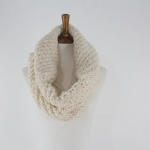 May include: Cream-colored, chunky knit infinity scarf displayed on a mannequin. The scarf is made of a thick, textured yarn, creating a warm and cozy appearance. The neutral color makes it a versatile accessory for various outfits.