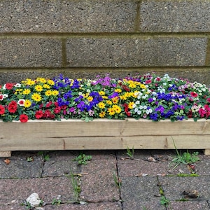 以下が含まれることがあります： 色とりどりの花が咲き誇る長方形の木製プランター。赤い花、黄色い花、青い花、白い花などがあります。プランターはレンガの表面に置かれ、灰色のレンガの壁を背景にしています。