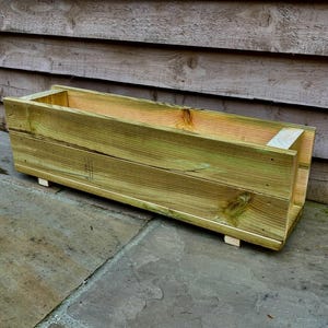 Könnte beinhalten: Rechteckiger Pflanzkasten aus hellgrün behandeltem Holz. Die Box ist mit sichtbarer Holzmaserung konstruiert und steht leicht erhöht auf kleinen Holzfüßen. Ideal für die Gartenarbeit im Freien.