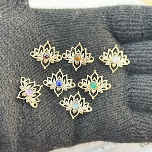 May include: Gold-colored lotus flower charms with various gemstone centers, including red, brown, blue, green, and purple. The charms are displayed on a dark gray surface.