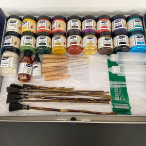 Op de afbeelding: Een kunstset met verschillende kleuren in potten, penselen, pipetten en flessen. De set bevat ook houten gereedschap en plastic flessen, alles netjes gerangschikt in een blauw-witte doos.