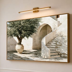 Op de afbeelding: Een rechthoekig wandkunstwerk in een gouden lijst met een olijfboom in een grote beige pot op een stenen terras naast een gebogen witte trap en een boogvormige ingang, verlicht door een horizontale gouden lamp erboven.