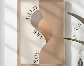 May include: A framed art print with a minimalist design. The artwork features abstract shapes in beige, tan, and gray tones, with the words "HELD ARE YOU" in black. The print is displayed on a white wall above a wooden bench.