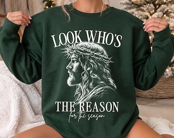 May include: A dark green crewneck sweatshirt featuring a white graphic of a profile view of Jesus wearing a crown of thorns. The text reads LOOK WHO'S THE REASON for the season in a mix of bold serif and script fonts centered on the chest.