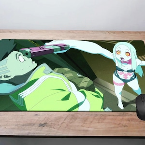 May include: A rectangular mouse mat with a colourful anime-style illustration. The artwork shows two characters in a dynamic scene with a gun. The colour palette includes shades of green, blue, and pink, with a black mouse resting on the mat.