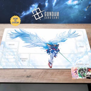 May include: A white Gundam card game playmat featuring a blue and white winged robot illustration. The mat has zones for cards and the text "Gundam Card Game". A few Gundam cards are visible in the lower right corner.