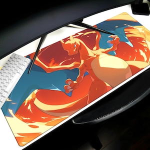 May include: A large rectangular mousepad featuring a vibrant illustration of a Charizard, a fiery orange and blue dragon-like creature. The mousepad is positioned on a white desk, with a white keyboard visible on the left.