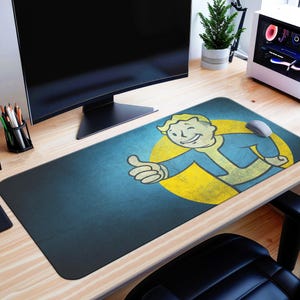 Puede incluir: Una alfombrilla de ratón rectangular con el personaje Vault Boy de la serie de videojuegos Fallout. El diseño incluye a Vault Boy haciendo una señal de pulgar hacia arriba, sobre un fondo azul y amarillo. La alfombrilla está sobre un escritorio de madera.