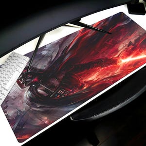 May include: A large rectangular gaming mouse pad with a dark fantasy art design. The artwork features a red and black color scheme with a central figure emitting a bright beam of light. A white keyboard and black monitor are visible.