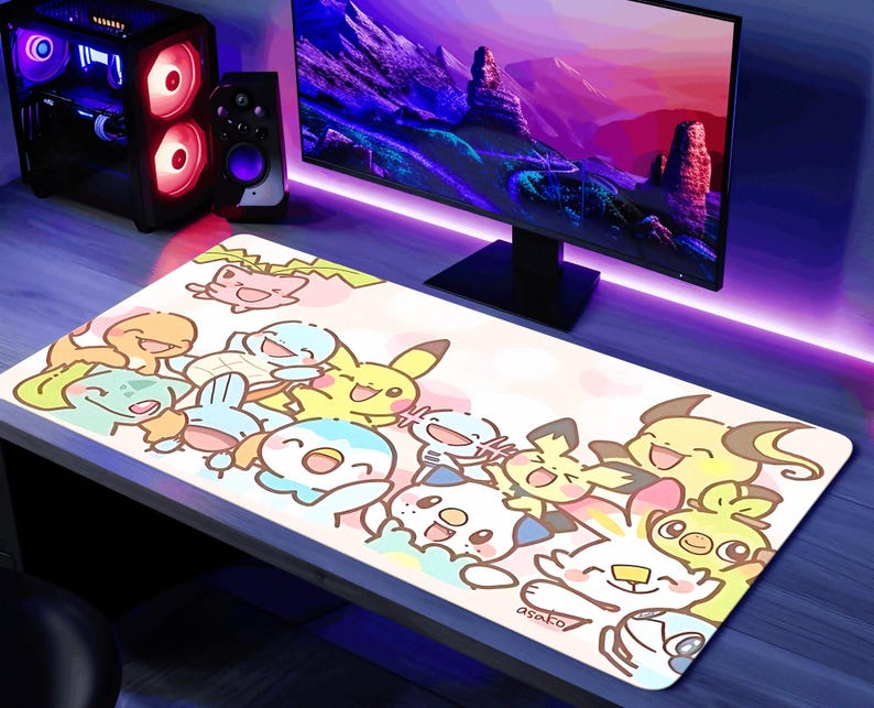 A gaming setup with a large monitor displaying a colorful mountain landscape, a keyboard with a mousepad featuring cartoon Pokemon characters, and a gaming console with a speaker and other peripherals.
