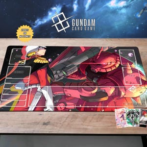 Może przedstawiać: Prostokątna mata do gry Gundam Card Game z czerwoną i czarną ilustracją robota i postaci Gundam. Mata ma strefy oznaczone do gry i tekst "Gundam Card Game". Widocznych jest kilka paczek kart.