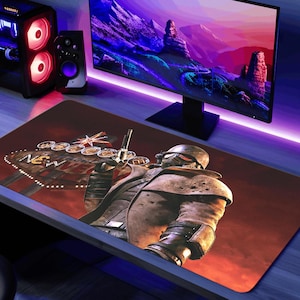 May include: A rectangular gaming mousepad featuring a detailed illustration of a character from the Fallout video game series, holding a gun. The design includes the "New Vegas" sign against a red and orange background. A computer monitor and tower are visible in the background.