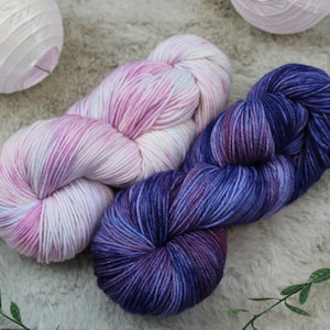 May include: Two skeins of colorful yarn. One is a blend of pink, white, and light blue, and the other is a deep purple with blue and red hues. The yarn is arranged on a soft, textured surface, suggesting a craft or knitting project.
