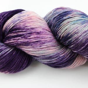 May include: A skein of hand-dyed yarn in shades of purple, pink, and blue. The yarn is tightly wound, showing a gradient of colors from deep violet to pale pink. The texture appears soft and suitable for knitting or crochet projects.