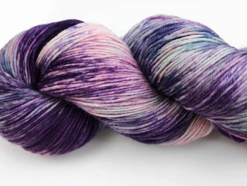 May include: A skein of hand-dyed yarn in shades of purple, pink, and blue. The yarn is tightly wound, showing a gradient of colors from deep violet to pale pink. The texture appears soft and suitable for knitting or crochet projects.