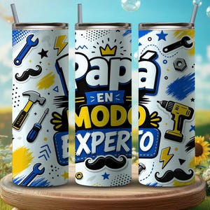 Puede incluir: Tres vasos aislados blancos con pajitas negras. Cada vaso presenta un diseño con herramientas, bigotes y el texto "Papá en Modo Experto" en azul y amarillo. Los vasos están sobre una superficie de madera.