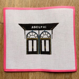May include: Needlepoint canvas featuring a black and gold architectural design with the word "ADELPHI" above two black doors. The canvas has a pink border and is on a white background.