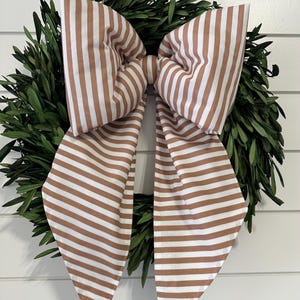 May include: A decorative wreath featuring a lush green base and a large bow. The bow is made of fabric with brown and white stripes. The wreath is displayed against a white background.