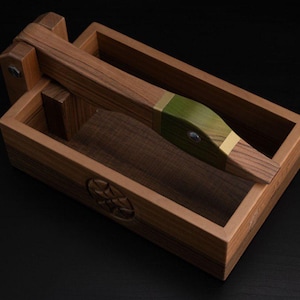 May include: A wooden nutcracker with a lever mechanism, resting in a rectangular box. The nutcracker features a green and gold accent on the head. The box has a carved emblem on the side.