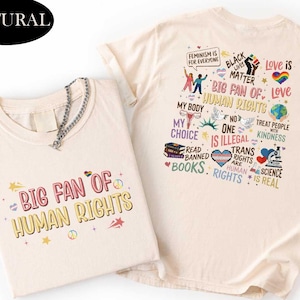 May include: Two cream-colored t-shirts with the text "BIG FAN OF HUMAN RIGHTS" in pink and yellow. One shirt has additional graphics and text including "Feminism is for everyone", "Black Lives Matter", and "Love is Love". A silver chain necklace is draped over one shirt.