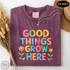 a purple t-shirt with the text "GOOD THINGS GROW HERE" printed on it, along with colorful flower illustrations. The t-shirt is laid out on a white surface, and there is a brown hat and some dried flowers in the background.
