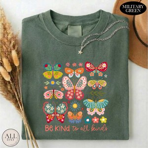 a green t-shirt with a colorful butterfly design and the text "Be Kind to all kinds" printed on it. The t-shirt is laid out on a white surface, with a brown hat and some dried flowers nearby.