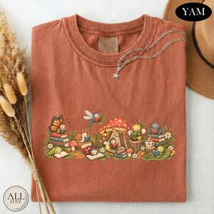 K&ouml;nnte beinhalten: Ein rostfarbenes T-Shirt mit einer grafischen Illustration verschiedener Insekten, darunter ein Schmetterling, eine Schnecke, ein Marienk&auml;fer und eine Biene, die unter einem gro&szlig;en Pilz B&uuml;cher lesen. Das Design zeigt zudem kleine Blumen.
