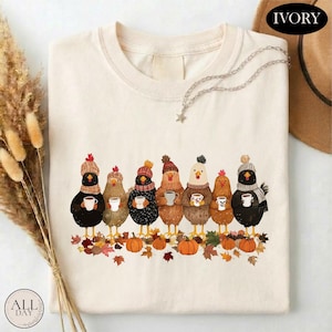 May include: An ivory t-shirt featuring a graphic of seven chickens wearing knit hats and scarves, each holding a mug. The chickens stand above a pile of colorful autumn leaves and pumpkins. The word IVORY is printed in the top right corner.