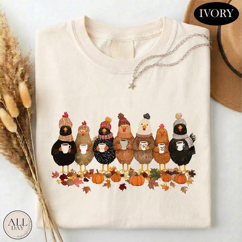 May include: An ivory t-shirt featuring a graphic of seven chickens wearing knit hats and scarves, each holding a mug. The chickens stand above a pile of colorful autumn leaves and pumpkins. The word IVORY is printed in the top right corner.