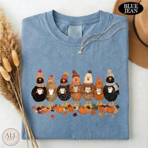 May include: A blue t-shirt featuring a graphic of seven chickens wearing knit hats and scarves, each holding a white mug. The chickens are standing among scattered autumn leaves and orange pumpkins. The shirt has a crew neckline and short sleeves.