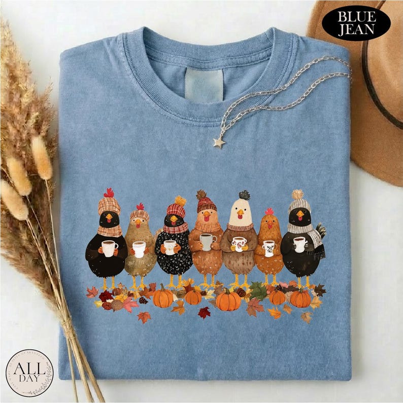 May include: A blue t-shirt featuring a graphic of seven chickens wearing knit hats and scarves, each holding a white mug. The chickens are standing among scattered autumn leaves and orange pumpkins. The shirt has a crew neckline and short sleeves.