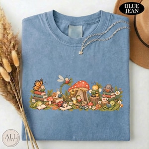 K&ouml;nnte beinhalten: Ein blaues T-Shirt mit einer grafischen Illustration verschiedener kleiner Kreaturen, darunter ein Schmetterling, eine Schnecke, ein Marienk&auml;fer und eine Biene, die inmitten von Pilzen und Laub B&uuml;cher lesen. Eine silberfarbene Kette mit Sternanh&auml;nger.