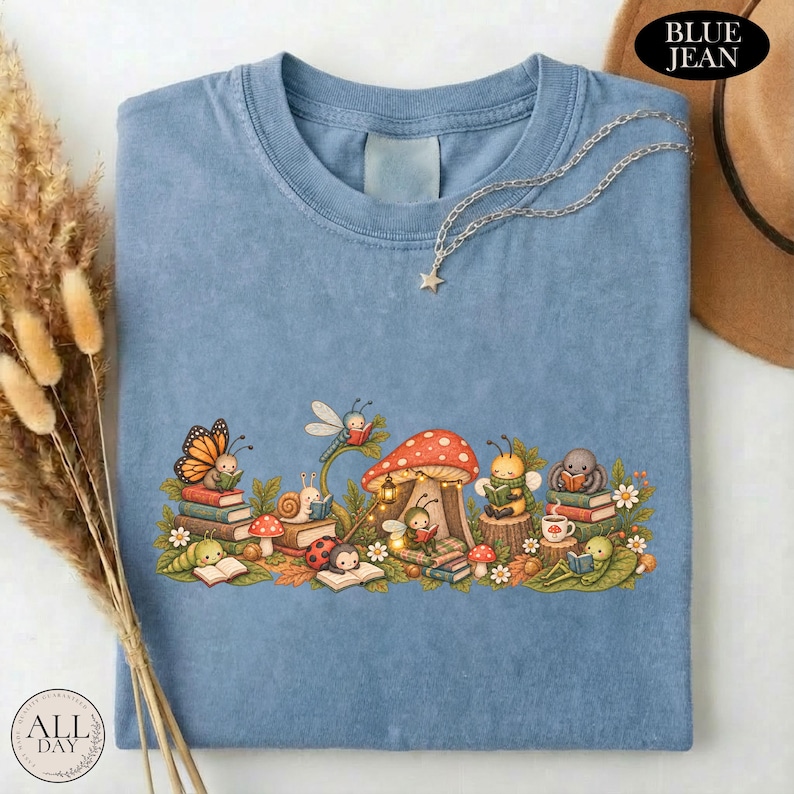 K&ouml;nnte beinhalten: Ein blaues T-Shirt mit einer grafischen Illustration verschiedener kleiner Kreaturen, darunter ein Schmetterling, eine Schnecke, ein Marienk&auml;fer und eine Biene, die inmitten von Pilzen und Laub B&uuml;cher lesen. Eine silberfarbene Kette mit Sternanh&auml;nger.