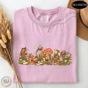 K&ouml;nnte beinhalten: Ein hellviolettes T-Shirt mit einer grafischen Illustration verschiedener Insekten, die um einen gro&szlig;en Pilz herum B&uuml;cher lesen. Das Design zeigt einen Schmetterling, eine Schnecke, einen Marienk&auml;fer und andere kleine Wesen inmitten von Laub.