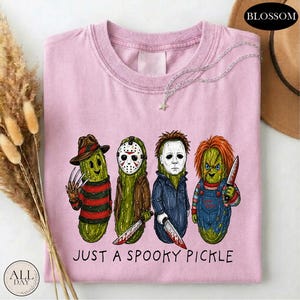 May include: A light pink t-shirt featuring a graphic of four green pickles dressed as horror movie characters. Below the pickles, the text reads "JUST A SPOOKY PICKLE". The shirt has a crew neckline and a short-sleeved design.