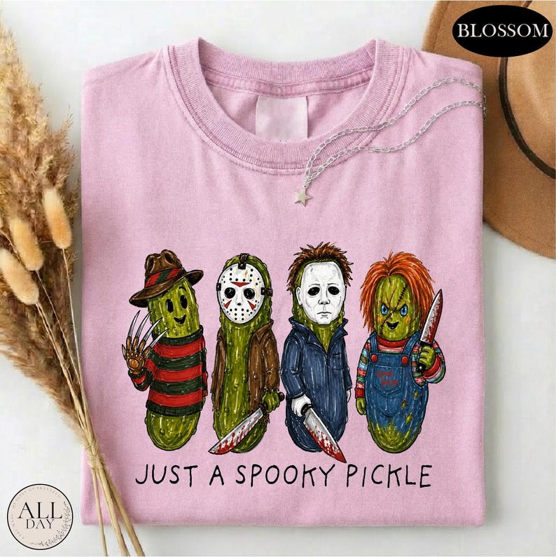 May include: A light pink t-shirt featuring a graphic of four green pickles dressed as horror movie characters. Below the pickles, the text reads "JUST A SPOOKY PICKLE". The shirt has a crew neckline and a short-sleeved design.