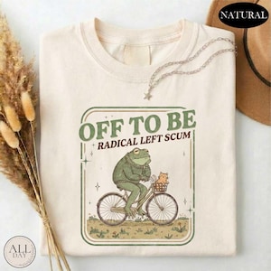 May include: Cream-colored t-shirt with a green and brown graphic. The graphic features a frog riding a bicycle with a cat in the basket, and the text "OFF TO BE + RADICAL LEFT SCUM". A silver necklace with a star pendant is draped on the shirt.
