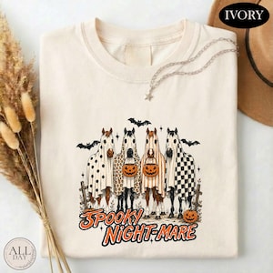 May include: A cream-colored t-shirt featuring a graphic of four horses wearing ghost costumes, each holding a jack-o'-lantern. The horses are decorated with polka dots, leopard print, stripes, and checkerboard patterns, with the text SPOOKY NIGHT-MARE.