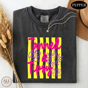 a black t-shirt with a colorful graphic design, including yellow and purple stripes and a sun-like symbol. The text "Tanned & Tipsy" is printed on the t-shirt.
