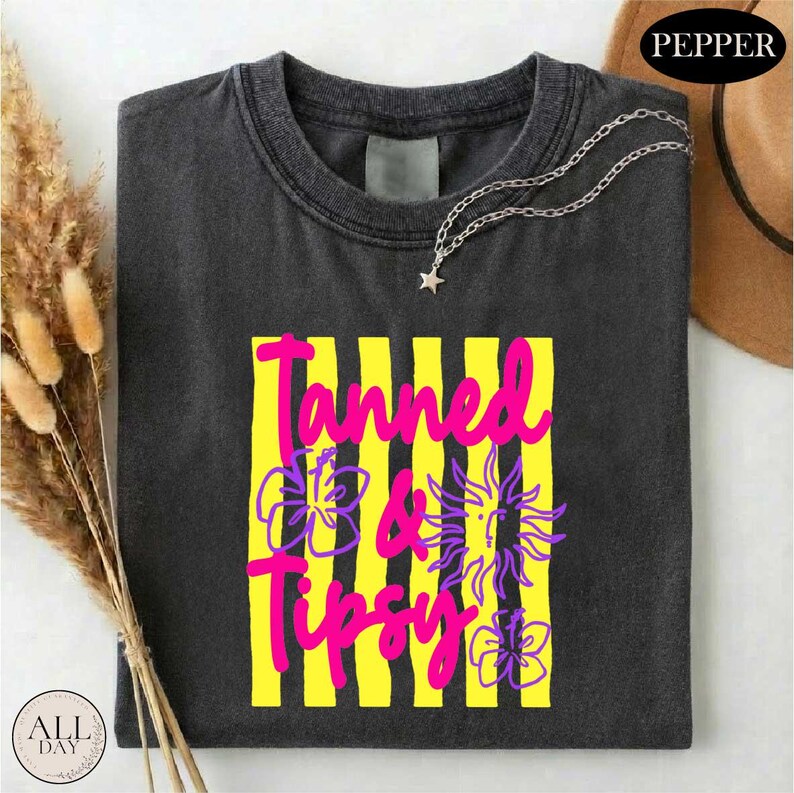 a black t-shirt with a colorful graphic design, including yellow and purple stripes and a sun-like symbol. The text "Tanned & Tipsy" is printed on the t-shirt.