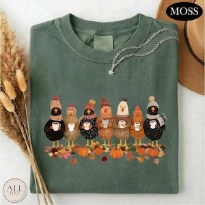 May include: A moss green t-shirt featuring a graphic of seven chickens wearing winter hats and scarves, each holding a white mug. The chickens stand above a pile of autumn leaves and orange pumpkins. A silver star necklace rests on the neckline.