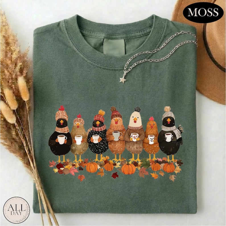 May include: A moss green t-shirt featuring a graphic of seven chickens wearing winter hats and scarves, each holding a white mug. The chickens stand above a pile of autumn leaves and orange pumpkins. A silver star necklace rests on the neckline.