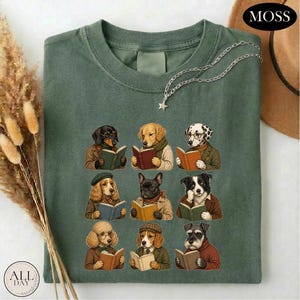 May include: A moss green t-shirt featuring a graphic print of nine different dog breeds, each wearing clothing and reading an open book. The dogs are arranged in a three-by-three grid, showcasing various breeds including a poodle and a dachshund.