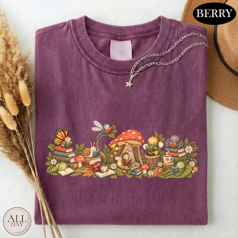 K&ouml;nnte beinhalten: Ein beerenfarbenes T-Shirt mit einer grafischen Illustration verschiedener Insekten, darunter ein Schmetterling, eine Schnecke, ein Marienk&auml;fer und eine Biene, die um ein Pilzhaus herum B&uuml;cher lesen. Eine silberne Sternkette liegt am Ausschnitt.