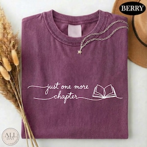 May include: A berry-colored crew neck t-shirt featuring the white text "just one more chapter" in a cursive font alongside a simple line-art illustration of an open book. A silver-toned chain necklace with a small star charm rests on the collar.