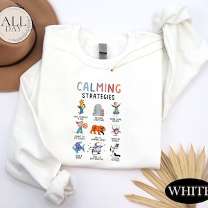 a white long-sleeved shirt with a colorful graphic design of various animals and objects, along with the text "Calming Strategies". The shirt is laid out on a white background, and there is a hat and a fan visible in the background.