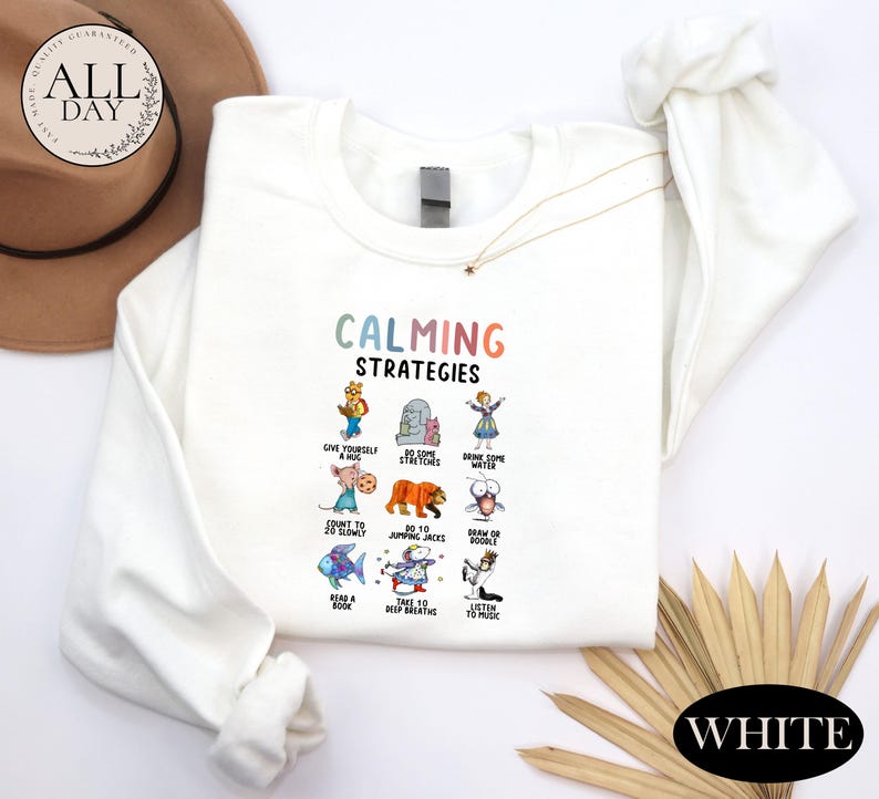 a white long-sleeved shirt with a colorful graphic design of various animals and objects, along with the text "Calming Strategies". The shirt is laid out on a white background, and there is a hat and a fan visible in the background.