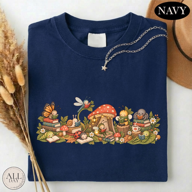 K&ouml;nnte beinhalten: Ein marineblaues T-Shirt mit einer grafischen Illustration verschiedener kleiner Insekten und Kreaturen, die um einen gro&szlig;en Pilz herum B&uuml;cher lesen. Das Design zeigt einen Schmetterling, eine Schnecke, einen Marienk&auml;fer, eine Biene und eine Spinne.